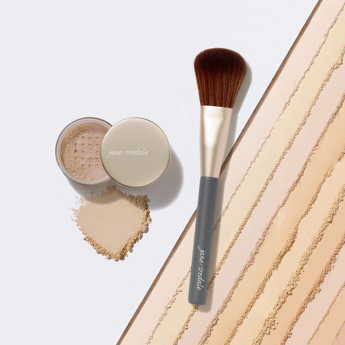 Geöffnete Dose des jane iredale Amazing Base Loose Mineral Powder neben einem Make-up-Pinsel. Daneben sind ein heller Puderauftrag und mehrere Pudernuancen auf weißem Hintergrund zu sehen.