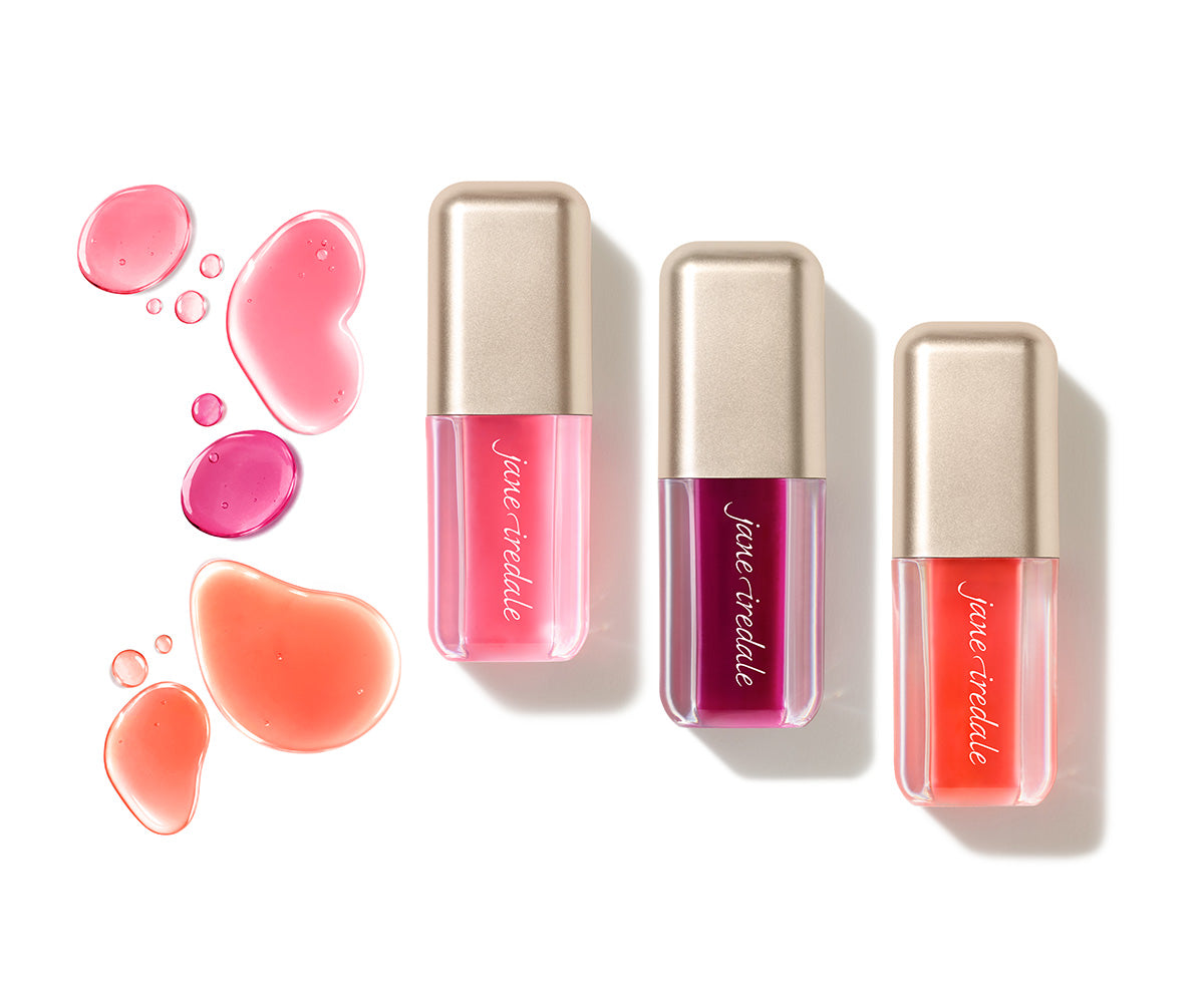 Drei jane iredale Just Kissed Lip Oils in Rosa, Beerenton und Koralle mit goldfarbenen Verschlüssen. Links daneben sind glänzende Tropfen der drei Farbnuancen auf weißem Hintergrund angeordnet.
