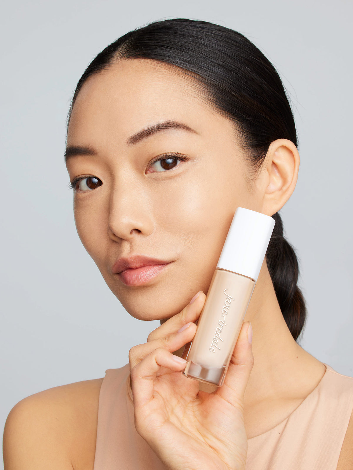 Ein weibliches Model mit schwarzen Haaren und braunen Augen hält die Glasflasche mit der jane iredale Flüssigfoundation Skintuition und schaut in die Kamera